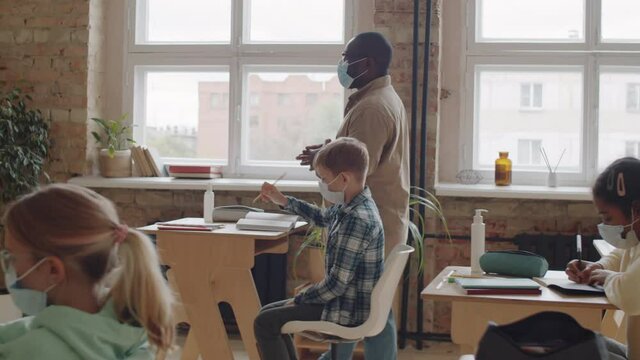 Afro-American male teacher in medical face mask walking through classroom and then standing by lectern and speaking while giving lesson to schoolchildren during coronavirus pandemic - Powered by Adobe