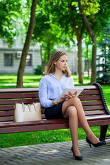 Obraz premium Young business woman in white shirt to go and touch pad during a break sitting outdoors in summer park on the background