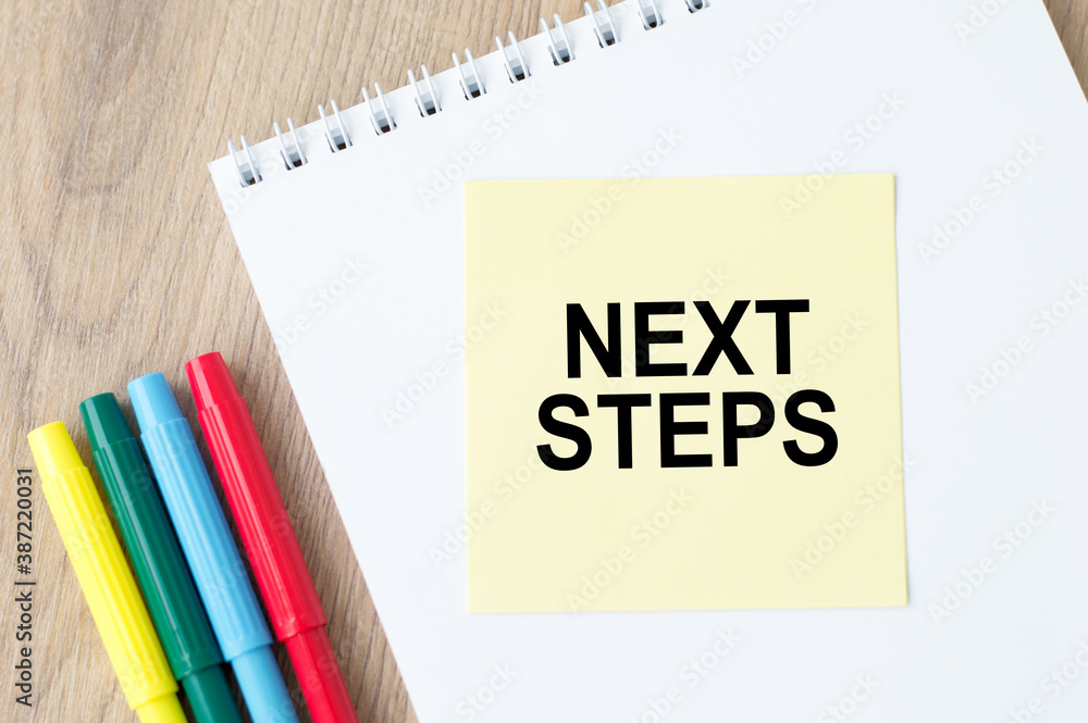 Poster next steps inscription on the sticker on an open notebook on the table next to colored markers