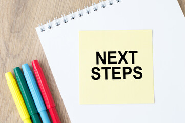 Next steps inscription on the sticker on an open notebook on the table next to colored markers