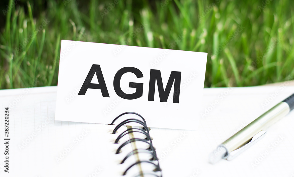 Poster the word agm on a card on an open notebook next to a pen on a green background.