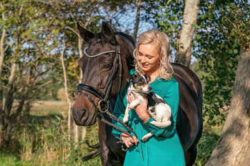 Beautiful portrait of a blond smiling girl with her horse and dog in the forest. Wearing a green dress. Selective focus