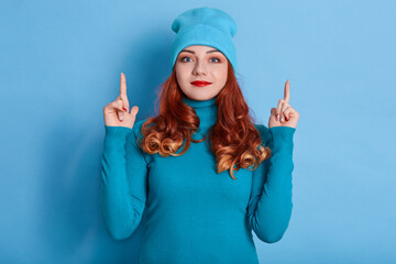 Close up portrait of clever red haired woman with curls, wearing cap, pointing fore fingers up,...