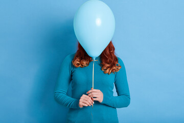 Portrait of woman hides her face behind helium balloon, red haired female with curls holding ballon...