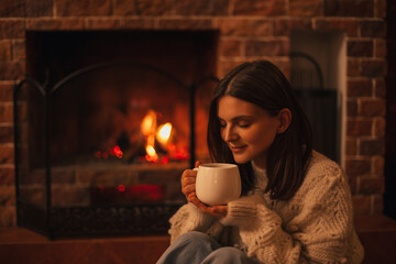 Young woman wearing white woolen knitted sweater enjoying hot tea near fireplace in a cozy living room. - obrazy, fototapety, plakaty