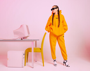 Stylish female office worker in sunglasses and hoodie standing with obsolete computer. Colored Neon light