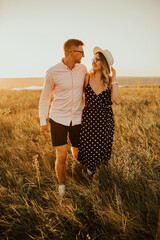 a guy with a girl in hat walking in the meadow. A couple of fair-haired fair-skinned people in love are resting in nature in a field at sunset.