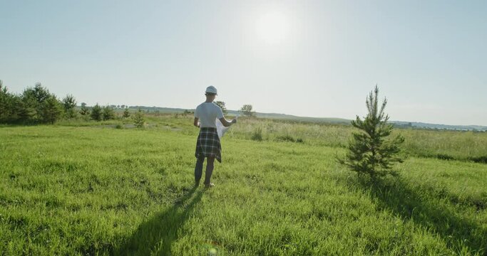 Male architect reading draft in field. Pan around view of man in hardhat unrolling and examining blueprint while standing in green grassy meadow in countryside