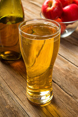 A glass of cold apple cider on an old rustic wooden table with a glass bottle and ripe red apples. Selected focus.