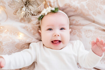 Close-up portrait of a little baby in a white dress with an ornament on her head. The girl lies on a light sofa in garlands of lights, a festive Christmas tree, laughing. Copy space, greeting card