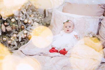 Close-up portrait of a little baby in a white dress with an ornament on her head. The girl lies on a light sofa in garlands of lights, a festive Christmas tree, laughing. Copy space, greeting card