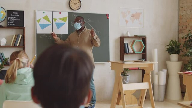 Black male teacher in protective face mask standing by chalkboard in front of children in classroom, pointing at posters and explaining math lesson while working in school during covid-19 outbreak