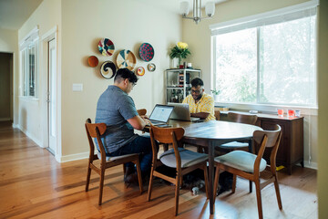 Gay couple working from home together on laptop computers sitting at dining room table 