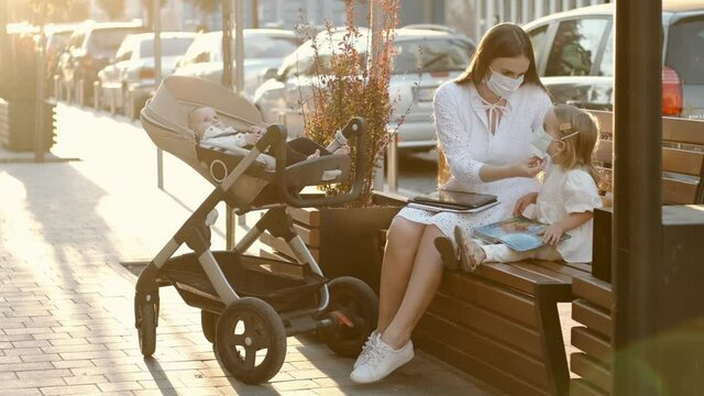 Maternity leave. Love and care. A mother is spending time with her kids outdoors. The woman is wearing mask and putting a mask on daughter's face. Mom is working while sitting on a bench with daughter