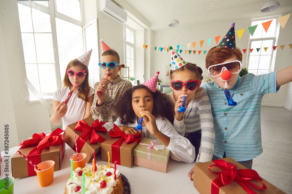 Wall mural Bunch of happy diverse kids in sunglasses and cone hats blowing noisemakers at birthday party