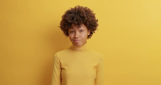 Young curly haired woman poses against yellow background has hesitant expression looks doubtful as asked difficult question decides something has confused perplexed face stands in studio