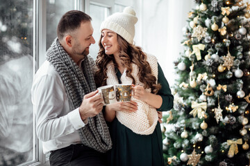 Christmas. Love. Home. Young couple in warm clothes is holding cups, talking and laughing while sitting at home near the Christmas tree
