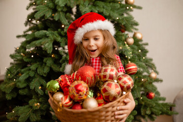 cute little girl in a santa hat holds a basket of toys against the background of the Christmas tree. Children dresses up christmas tree for new year