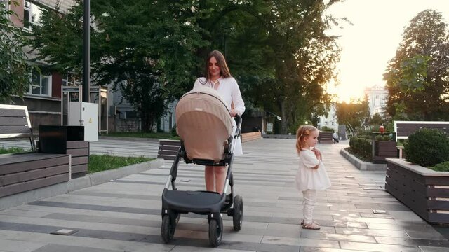 Caring mother is walking with a baby in a pram and taking a hand of a little daughter. The woman is taking care of children and giving her love. Family time,relaxing.