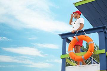 Male lifeguard on watch tower against blue sky - obrazy, fototapety, plakaty
