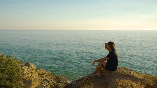 Hombre sentado en un acantilado al atardecer con el mar, el horizonte y el cielo de fondo, mirando, se&ntilde;alando con el dedo y saludando. Desde Conil, C&aacute;diz, Andaluc&iacute;a, Espa&ntilde;a, Europa.