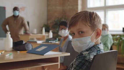 Selective focus shot of little caucasian boy in medical face mask sitting at desk in classroom and looking at camera while posing during school lesson
