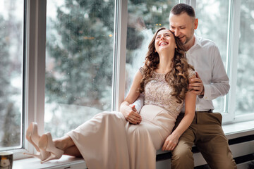 couple in love in festive clothes sitting and hugging near the large window and Christmas tree in decorated studio. happy holiday in winter time