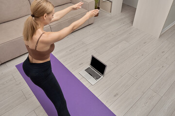 Back view of a young woman looking at a blank laptop screen, learning yoga poses online. Playing sports at home.