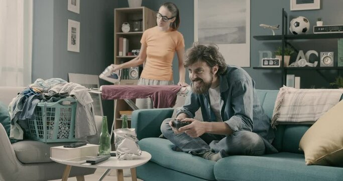 Lazy husband playing videogames while his wife is ironing