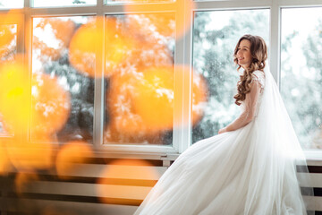 Fototapeta premium Luxury bride in wedding dress for Christmas near the large panoramic window. Luxury bride in wedding dress for Christmas. Charming bride in an elegant wedding dress. Bride in luxurious suites.