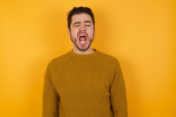 Young man wearing casual sweater and over isolated yellow background yawns with opened mouth stands. Daily morning routine