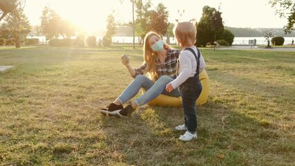 Loving mother in mask seated on beanbag chair embracing her cute toddler daughter, bonding and relaxing in summer. Family is in a park. The parent trying to put a mask on daughter's face. Disappointed - Powered by Adobe