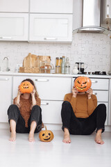 Young couple man and woman sitting on the floor at kitchen at home holding colorful pumpkins near their faces and preparing for halloween talking laughing cheerful.