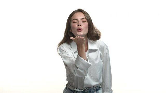 Video of beautiful romantic woman in white shirt, sending her love with air kiss and smiling, standing against studio background