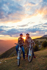 Fototapeta na wymiar Elderly man and woman on e-bikes holding their thumbs up on mountain. Beautiful sunset at background.