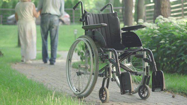 Wheelchair standing on park alley in sunlight with blurred senior Caucasian couple walking away at the background. Old husband and wife strolling outdoors with weak spouse on summer day.