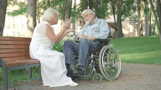 Wide shot of positive smiling husband and wife using smartphone in sunny park. Loving senior Caucasian woman sitting on bench with disabled husband in wheelchair and talking.