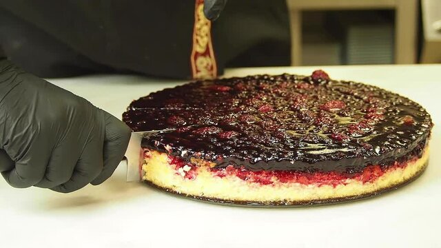 Professional chef is cuts off a piece of cheesecake decorated with fresh berries with a knife on a white kitchen table. The face is not visible.