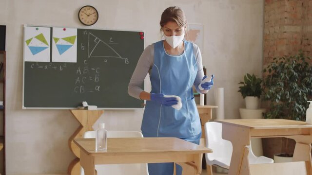 Young cleaning lady in uniform, protective face mask and gloves using sanitizing spray and napkin while disinfecting school desks in classroom during coronavirus outbreak