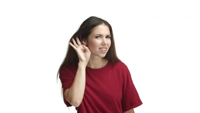 Pretty brunette woman in red t-shirt looking confused, cannot hear you, put hand near ear and asking to repeat question, standing over white background