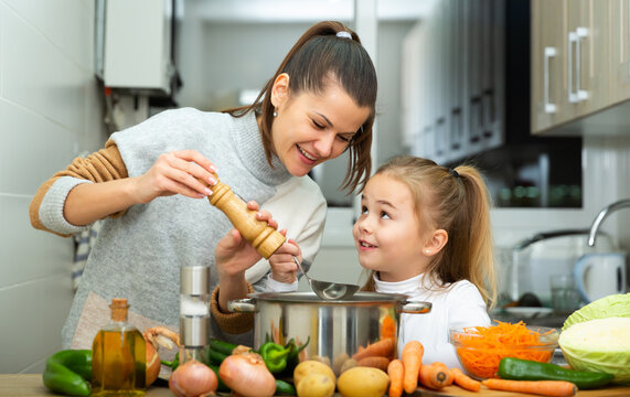Little daughter helping cooking soup and mother add pepper to pan