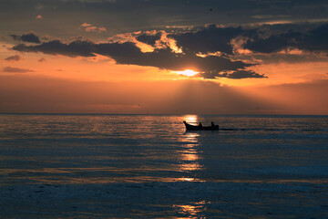 Naklejka na ściany i meble sunset over the sea with a small fishing boat and two fishermans over water and front the sun line ; sunlight within clouds : location : martil morocco tetouan mediterranean sea 