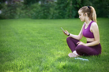 Young Smiling Blonde Athletic Girl Sitting On Grass In A Park With Smartphone And Earphones