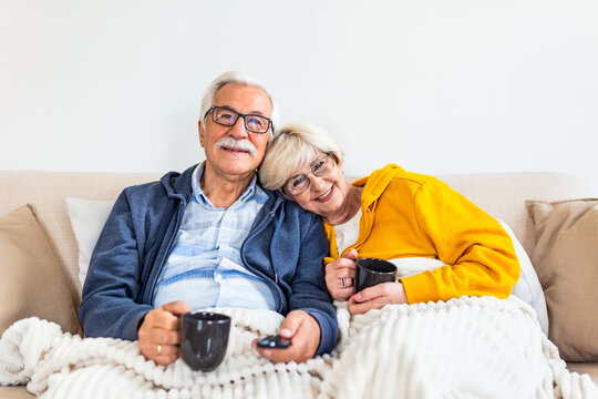 Happy Senior Couple Embracing And Watching TV, Sitting On Sofa In Living Room drinking hot tea and getting cozy under the blanket