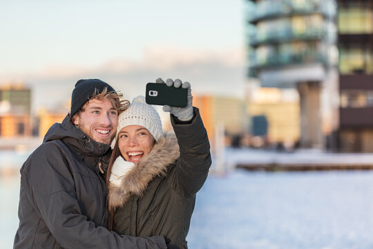 Selfie couple happy winter travel walk tourists taking photo with phone on city street panorama lifestyle. Asian woman, Caucasian man wearing knit hats and jackets.