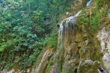 The stream flows down the mountainside and forms a small waterfall with thin streams. Lush green vegetation around.