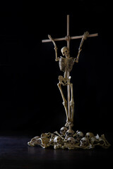 Skeleton tied up with crucifix wich has pile of skulls and bone below all put on dark background