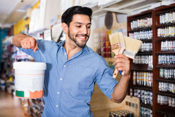 Satisfied young smiling man standing amongst racks in paint store with brushes and paint