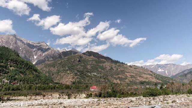 Timelapse of a house partially hidden by forest with the Himalayan Mountains in the background near Manali, Himachal Pradesh, India. Clouds drift across the mountain peaks in a tranquil alpine landsca