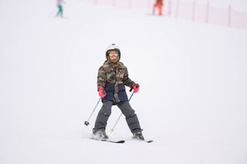 active little sport boy in helmet and khaki ski suit riding skiing with poles on winter white snow slope